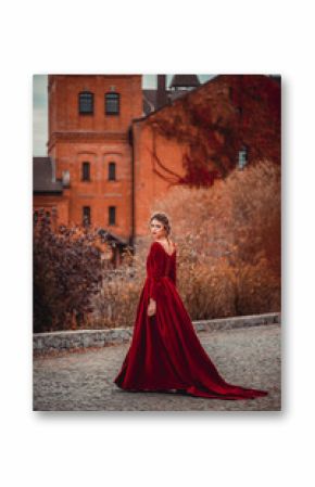 Beautiful girl in a burgundy red dress walking near  old castle on a background of autumn grape leaves in the park, October. Radomyshl