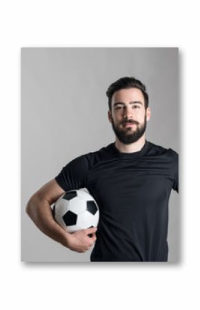 Friendly smiling bearded soccer player holding ball under his arm looking at camera over gray studio background.