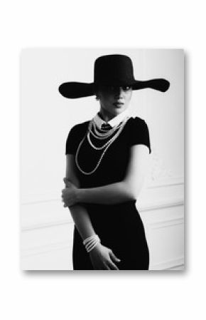 Elegant beautiful young woman in retro style dress, hat standing over light grey background. Black and white monochrome photo