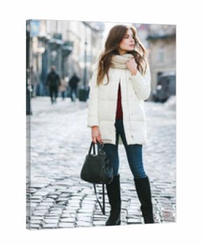 Outdoor full body portrait of young beautiful fashionable woman wearing stylish white winter puffer coat, suede high boots, holding bag. Model posing in street of the city. Female fashion concept
