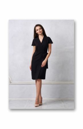 Attractive woman with long brunette hair dressed in fashionable black wrap around midi dress with short sleeves smiling and posing. Laughing female model standing against white wall on background.