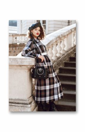 Outdoor full body fashion portrait of young beautiful fashionable girl wearing autumn long checked dress, leather beret, boots, holding black bag, posing in european city, near old architecture