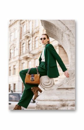 Outdoor full body fashion portrait of  fashionable woman wearing sunglasses, white turtleneck, green suit, blazer, trousers, leopard print boots, holding suede bag, posing in street of european city