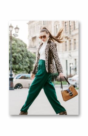 Outdoor full body fashion portrait of  fashionable woman wearing sunglasses, white turtleneck, leopard print blazer, boots, green trousers, holding brown suede bag, walking in street of european city