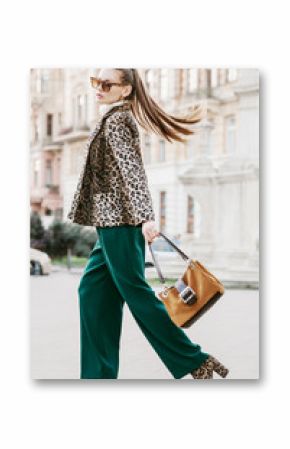 Outdoor full body fashion portrait of young beautiful fashionable woman wearing sunglasses, leopard print blazer, boots, green trousers, holding brown suede bag, walking in street of european city