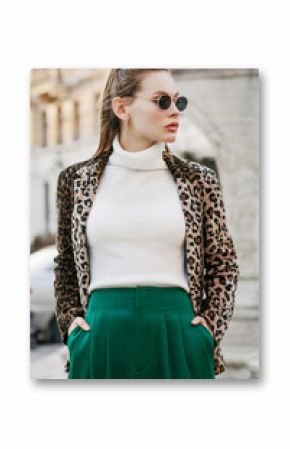 Outdoor fashion portrait of young beautiful confident woman wearing trendy leopard print blazer, white turtleneck, green high-waisted trousers, stylish sunglasses, posing in street of european city