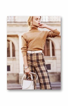 Outdoor fashion portrait of young beautiful fashionable girl wearing trendy beige cashmere turtleneck, high-waisted checkered trousers, belt, wrist watch, holding small white bag, posing in street