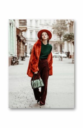 Outdoor full body fashion portrait of young beautiful woman wearing trendy oversized orange faux fur coat, hat, green sweater, corduroy trousers, holding stylish snakeskin bag, posing in street 