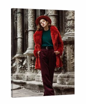 Outdoor full body fashion portrait of young beautiful woman wearing trendy oversized orange faux fur coat, hat, green sweater, stylish high-waisted corduroy trousers, posing in street of european city
