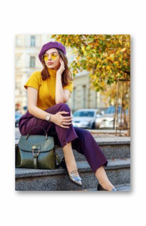 Outdoor full body fashion portrait of young elegant woman wearing purple beret, trousers, yellow glasses, turtleneck, loafers, wrist watch, with green leather handbag. Autumn street style concept