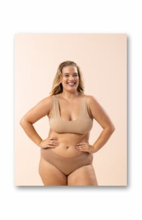 A young Caucasian plus-size model in beige underwear stands on beige background, copy space