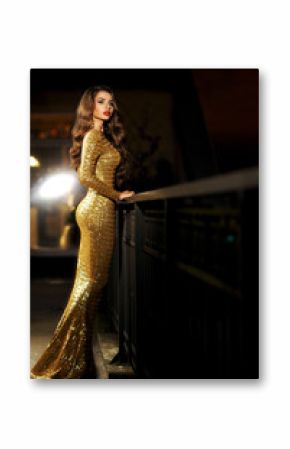 Sexy fashionable beautiful young woman in long golden shining evening dress standing at balcony over view on night city lights in bokeh. Pretty lady with long curly hair and red lips