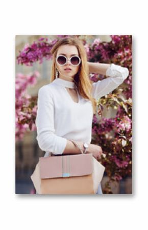 Outdoor waist up portrait of young beautiful girl posing in street. Model wearing stylish round sunglasses, white shirt, wrist watch, holding pink bag, handbag. City lifestyle. Female fashion concept