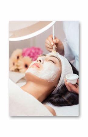 Close-up of the face of a young woman relaxing under the gentle touch of the specialist applying on her cheeks, white facial mask with rejuvenating effects