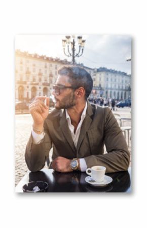 Handsome young man drinking espresso coffee and smoking cigarette, wearing elegant coat posing at table outside on urban background.
