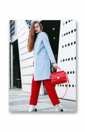 Outdoor full body portrait of young beautiful fashionable woman walking in street. Model wearing light blue coat, red pants, silvery ankle boots, holding trapeze handbag. Female fashion concept