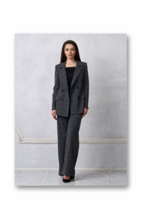 Gorgeous young woman dressed in gray squared jumpsuit, blazer and heeled shoes posing in studio. Beautiful brunette girl demonstrating stylish smart clothing against white wall on background.