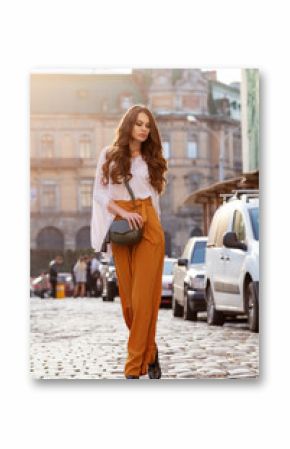 Outdoor full body fashion portrait of young beautiful woman wearing stylish yellow high-waisted wide-leg pants, white blouse, holding green leather bag. Model walking in street of european city