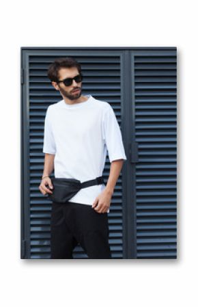 Fanny pack, bum bag. Men's street youth fashion. Young hipster man in sunglasses with a beard posing in the street on a background of roller shades blinds. White T-shirt and black shorts.