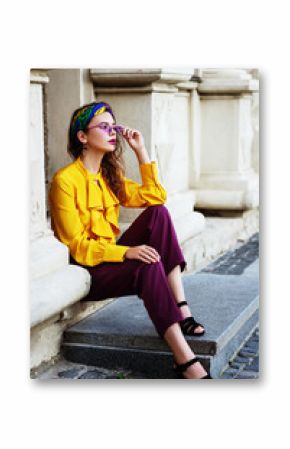Outtdoor full body fashion portrait of young beautiful confident woman wearing stylish headband, yellow blouse, violet trousers, trendy sunglasses, posing in street of european city.