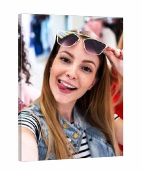 Smiling girlfriends wearing stylish sunglasses having fun time taking selfie with mobile phone while doing shopping in clothing store