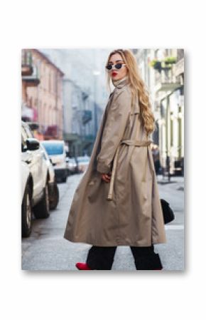 Outdoor full body fashion portrait of young beautiful fashionable woman wearing trendy beige long trench coat, stylish sunglasses, black trousers, red boots, model walking in street of european city