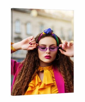Outdoor close up fashion portrait of young beautiful woman wearing trendy violet sunglasses, colorful headband, circle hoop earrings, yellow blouse, pink blazer, posing in street of european city. 