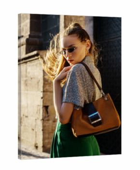 Outdoor fashion portrait of young beautiful fashionable woman walking street, model wearing sunglasses, leopard print shirt, green trousers, holding brown suede bag
