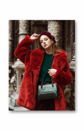 Outdoor fashion portrait of young beautiful confident woman wearing trendy orange faux fur coat, beret, green sweater, corduroy trousers, holding stylish snakeskin handbag, posing in street of city