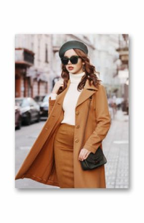 Outdoor portrait of young beautiful fashionable woman wearing trendy autumn coat, turtleneck, skirt, beret, sunglasses, with small shoulder bag, walking in street of european city