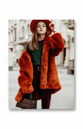 Outdoor fashion portrait of young beautiful confident woman wearing trendy orange faux fur coat, hat, green sweater, corduroy trousers, holding stylish handbag, posing in street of european city