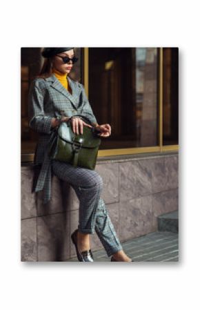 Outdoor full body fashion portrait of  elegant businesswoman, woman wearing glasses, beret, checked suite, blazer, trousers, loafers, holding leather handbag, posing in street of european city