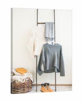 Fall or winter warm knitwear on hanger, white wall background. Fashionable female winter clothing. Woolen sweaters, grey scarf, boots and knitted hat