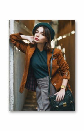 Outdoor fashion portrait of young woman wearing trendy corduroy blazer, green beret, checkered trousers, golden wrist watch, carrying leather bag,  posing in street of European city