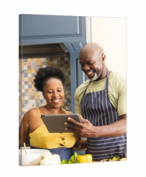 Happy senior african american couple preparing meal together, using tablet in kitchen