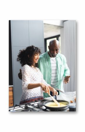 Happy mature diverse couple making breakfast, cooking eggs and talking in sunny kitchen, copy space