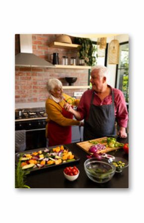 Happy caucasian senior couple putting on aprons, preparing vegetables in kitchen, copy space