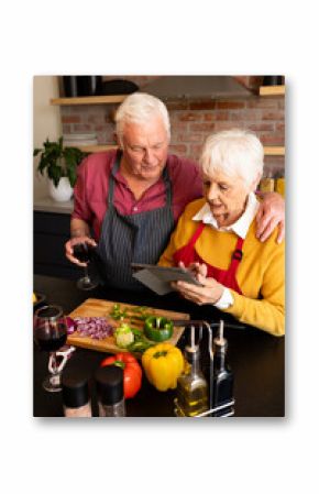 Happy caucasian senior couple preparing vegetables, drinking wine and using tablet in kitchen