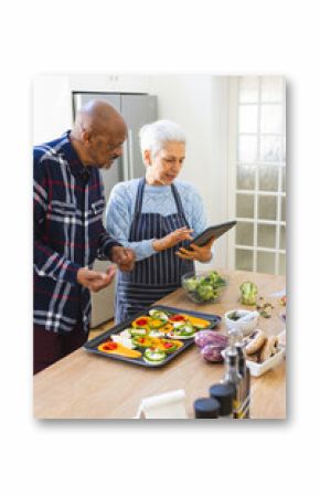 Diverse senior couple preparing meal using tablet in kitchen