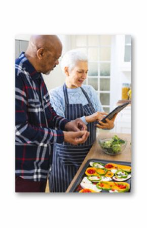 Diverse senior couple preparing healthy meal with vegetables using tablet in kitchen