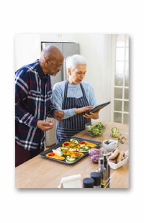 Diverse senior couple preparing healthy meal with vegetables using tablet in kitchen