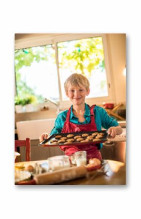 A eight years old blond boy with a red apron is cooking cookies