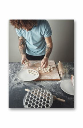 Tattooed curly chief man cooks pelmeni or ravioli in special mold. Collecting dropped dumplings in hand and on ceramic plate on rustic table, top view