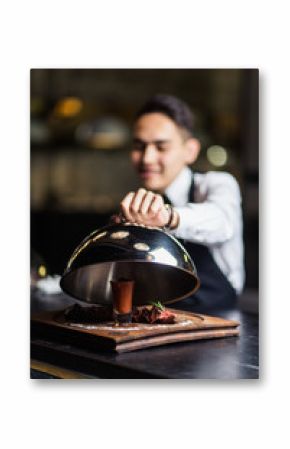 Attractive young waiter in tuxedo holding serving tray with metal cloche and napkin