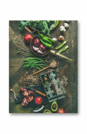 Winter vegetarian food cooking ingredients. Flat-lay of seasonal vegetables and fruit, beans, cereals, kitchen utencils, dried flowers, olive oil over wooden background, top view, vertical composition