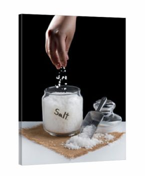 Hand sprinkling salt into glass jar over black background.