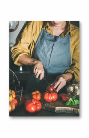 Woman in grey linen apron cutting and cooking tomato sauce, canned tomatoes or pasta with heirloom tomatoes, basil and garlic at kitchen counter. Seasonal healthy cooking, slow food or comfort food