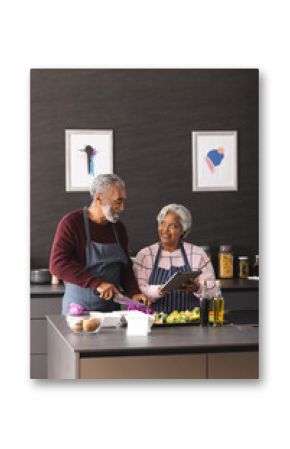 Happy senior biracial couple using tablet preparing vegetables in kitchen at home, copy space
