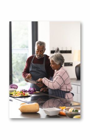 Happy senior biracial couple using tablet preparing vegetables in kitchen at home, copy space