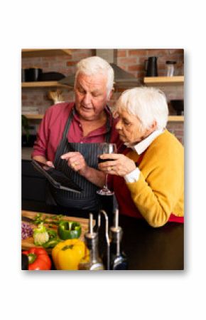 Happy caucasian senior couple preparing vegetables, drinking wine and using tablet in kitchen